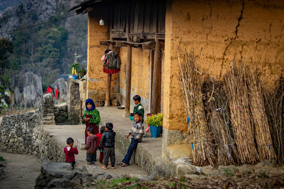 Children gather outside a mud-walled house in a rural village in Ha Giang, with limestone mountains behind.