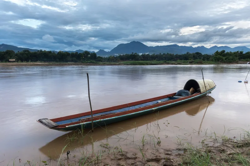 A wooden pirogue is moored on the muddy Mekong riverbank near Luang Prabang with mountains in the distance.