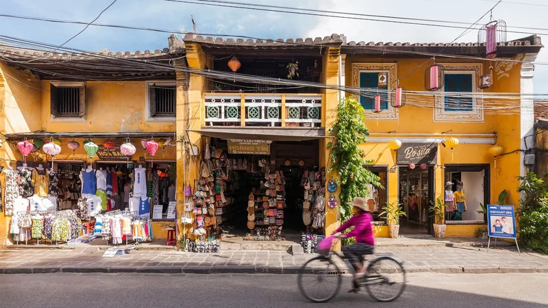 A cyclist passes yellow shophouses with lanterns and clothing stalls in Hoi An's old town.