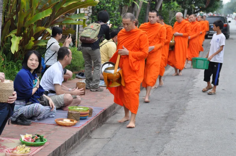 Barefoot monks in orange robes walk in line collecting alms from kneeling locals and tourists on a street in Luang Prabang.