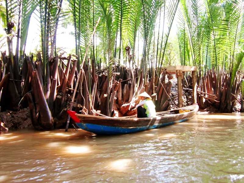 A woman in a conical hat paddles a small boat along a muddy Mekong Delta canal lined with water palms.