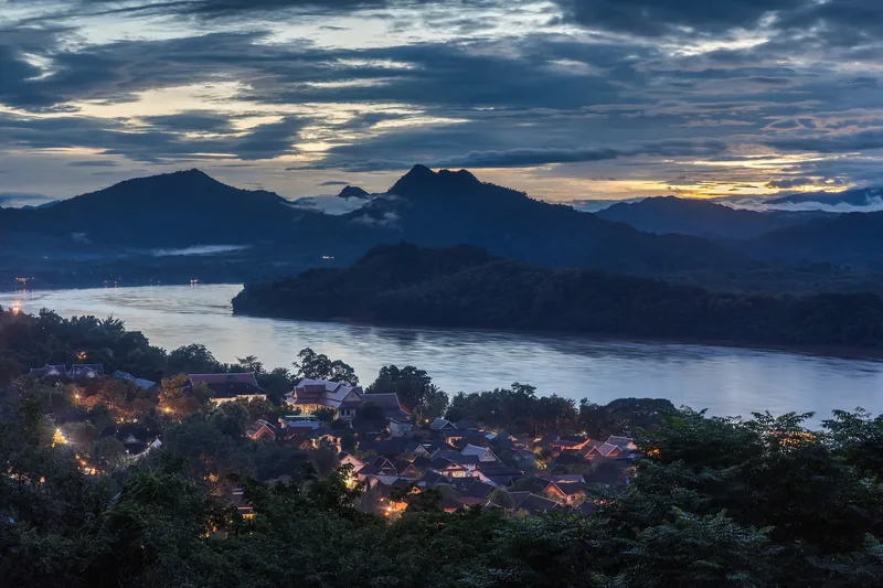 Twilight view over Luang Prabang's rooftops and the Mekong River winding between forested mountains.