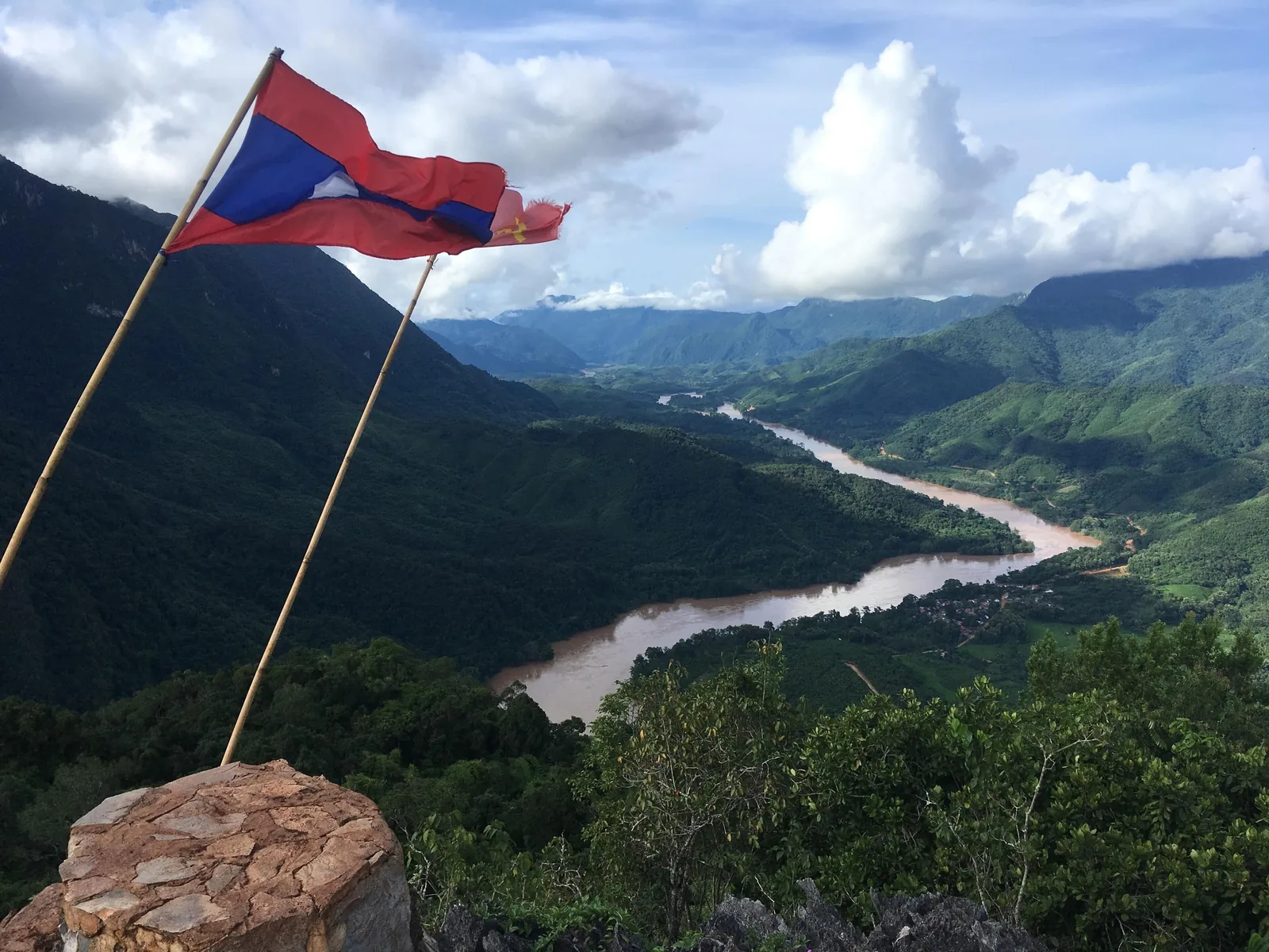 A Lao flag flutters on a stone cairn above the Nam Ou River winding through green mountains near Nong Khiaw.