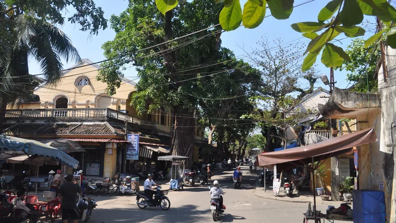 Motorbikes travel a shaded street lined with old yellow shophouses in Hoi An Ancient Town, Vietnam.