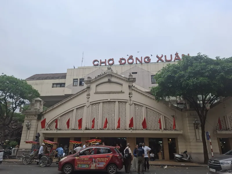 The historic arched facade of Dong Xuan Market with red flags and a large sign in Hanoi's Old Quarter.