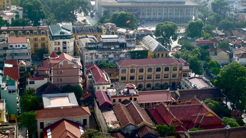 Aerial view of Hanoi's old quarter with red-tiled roofs, colonial buildings, and a hall visible in the background.