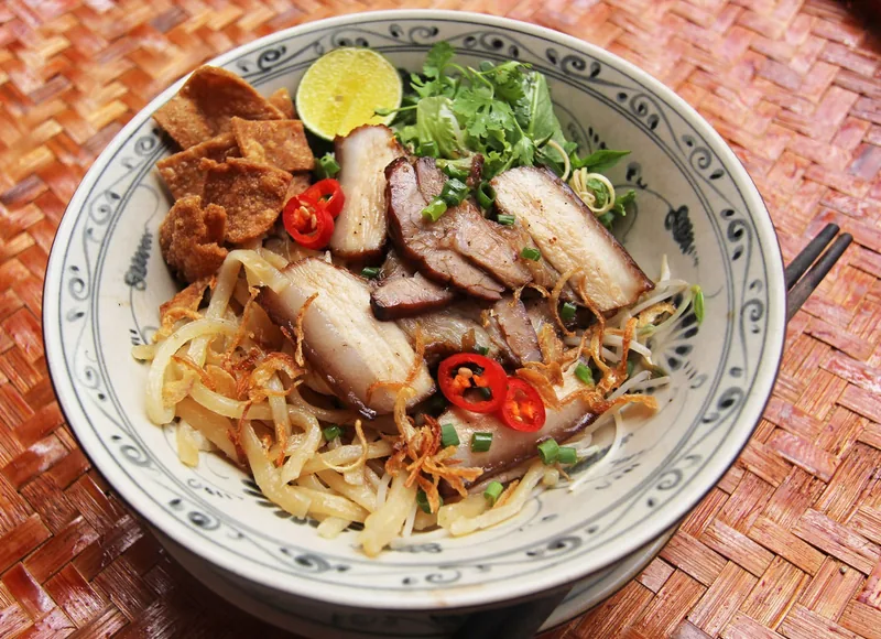 A bowl of Hoi An cao lau noodles topped with sliced pork, crispy crackers, herbs, chili and lime.