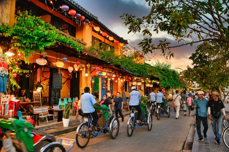 Colorful lanterns hang above cyclos and pedestrians on a lively street in Hoi An Ancient Town at dusk.