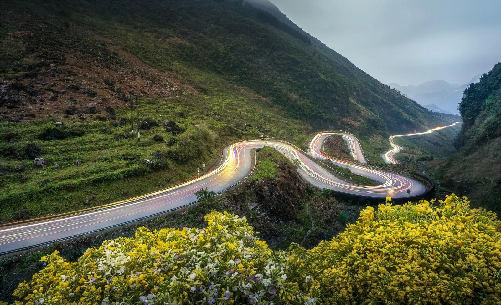 Light trails trace a winding mountain road through green hills and yellow flowers in Ha Giang.