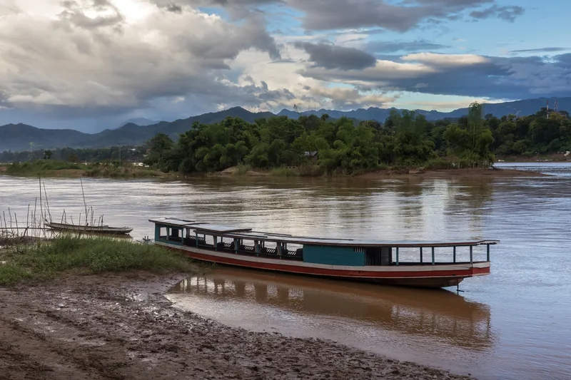 A long wooden boat rests on the muddy bank of the Mekong River in Luang Prabang under a cloudy sky.