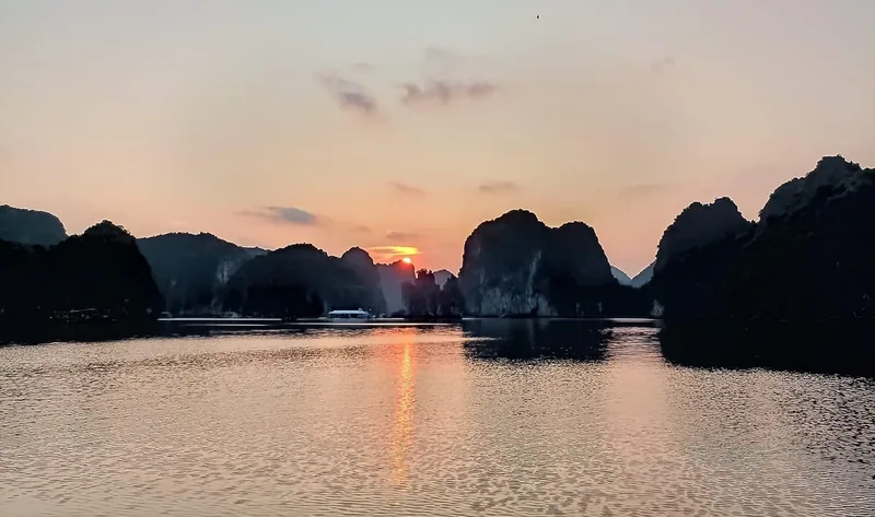 Sun sets between limestone karsts, casting a golden reflection over Lan Ha Bay's calm water.