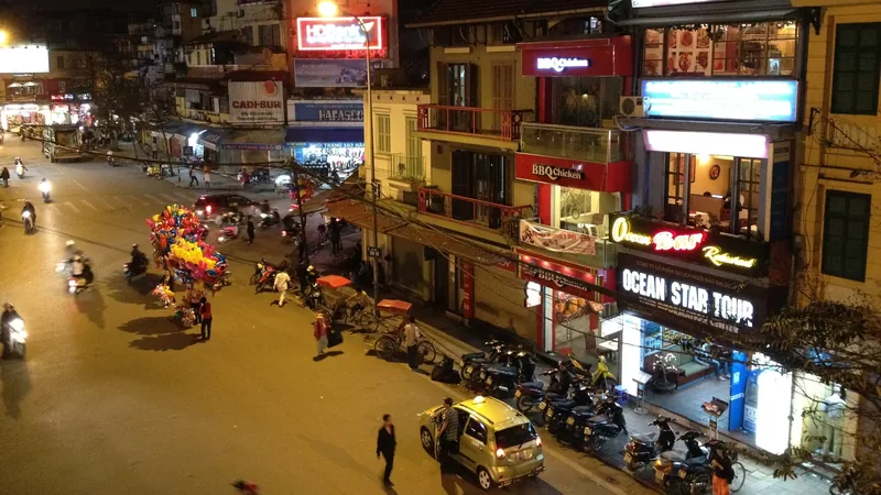 Aerial night view of a busy street corner in Hanoi's Old Quarter with neon signs, motorbikes and a taxi.