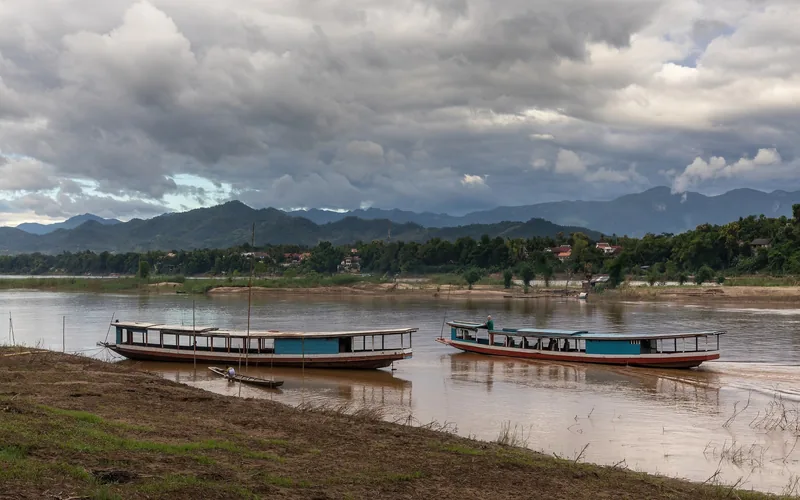 Two long wooden boats rest on the Mekong River near Luang Prabang under a cloudy sky and distant mountains.