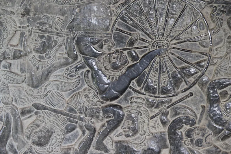 Detailed stone bas-relief carving depicting a chariot wheel and warrior figures at Angkor Wat in Siem Reap.