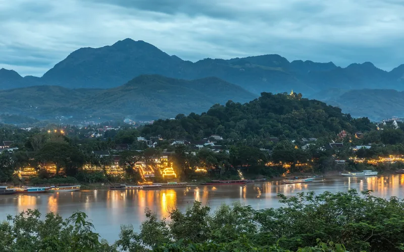 Twilight view over the Mekong River in Luang Prabang, with Mount Phousi and forested hills glowing under a golden riverside.