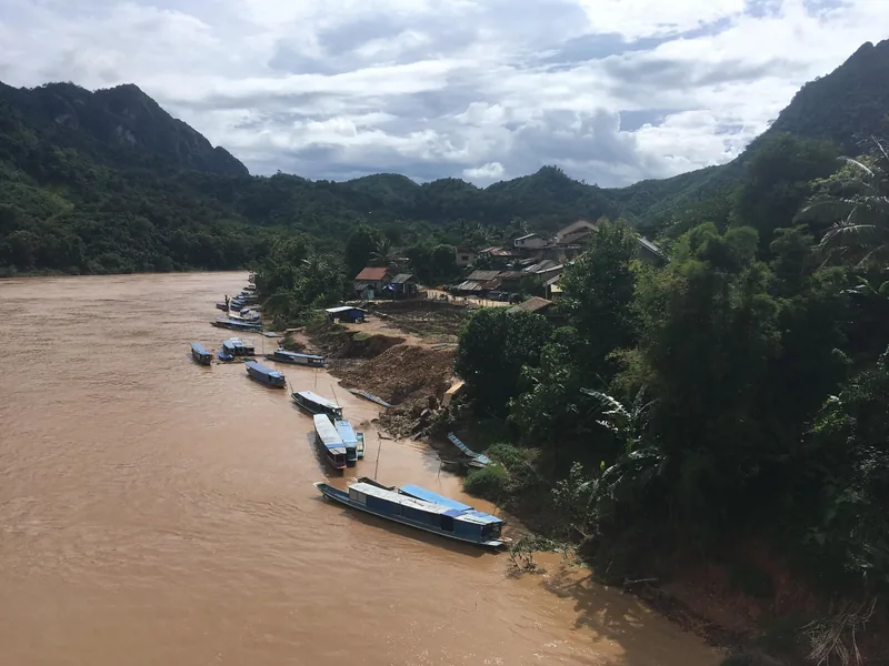 Wooden boats line the muddy Nam Ou River beside a riverside village in Nong Khiaw, framed by limestone mountains.