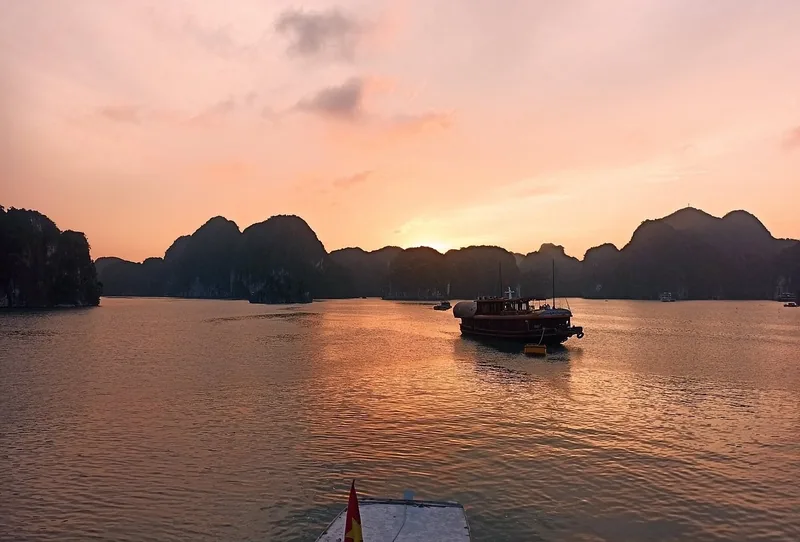 A wooden boat drifts across Lan Ha Bay as karst islands silhouette against an orange sunset sky.
