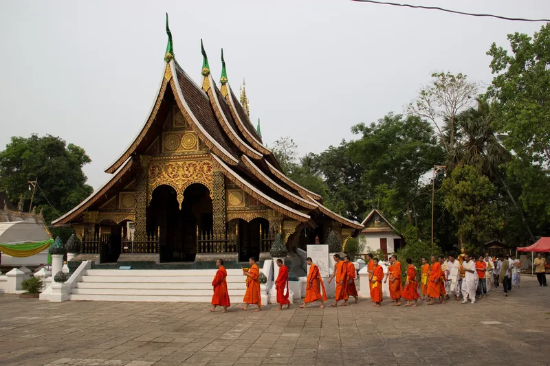 Buddhist monks in saffron robes walk in procession past a golden-roofed temple in Luang Prabang.