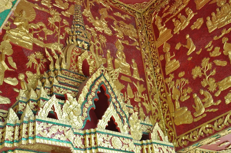 Gilded relief panels depicting Buddhist scenes decorate the red temple wall and roof gable in Luang Prabang.