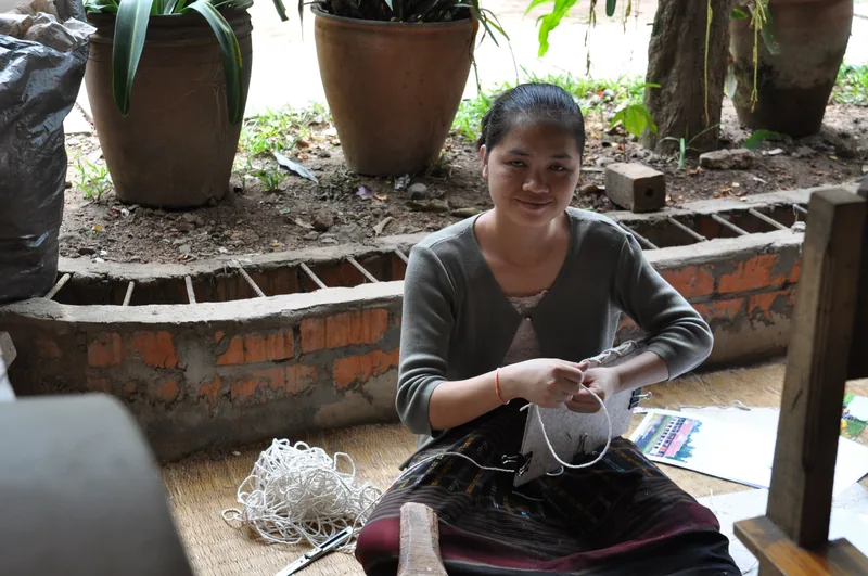 A woman in Luang Prabang sits smiling while handcrafting a bag from handmade paper cord.