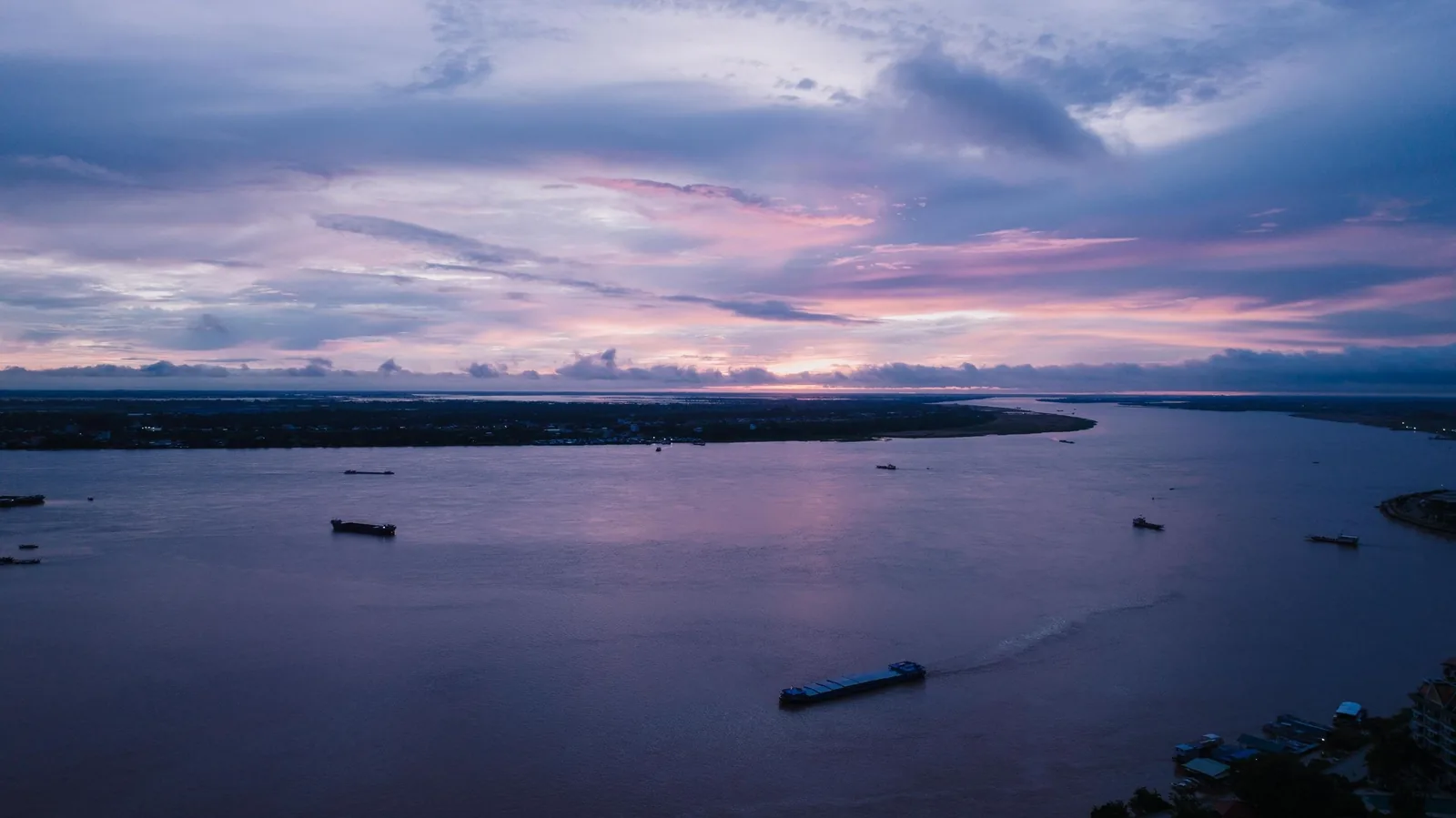 A pastel sunset sky glows over the wide Mekong River with cargo boats drifting near the delta shoreline.