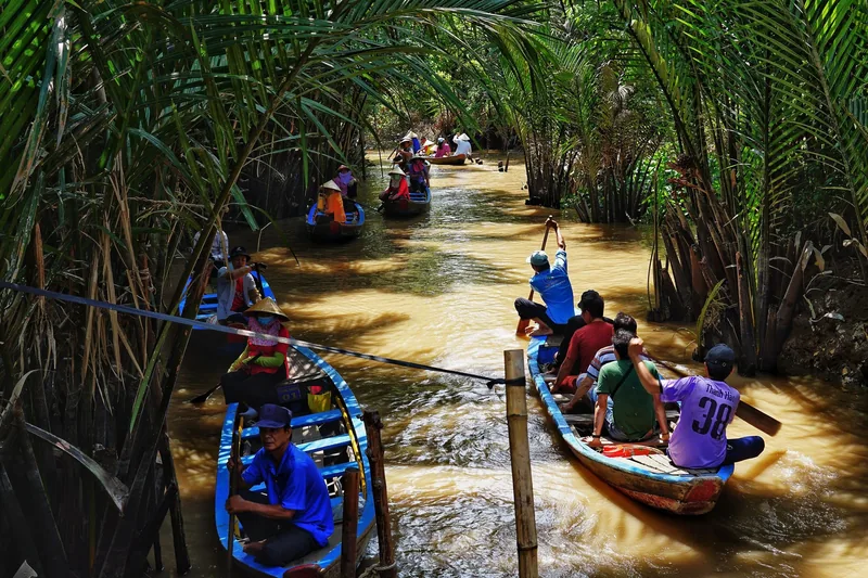 Wooden boats carry tourists along a narrow water channel shaded by palm fronds in the Mekong Delta.