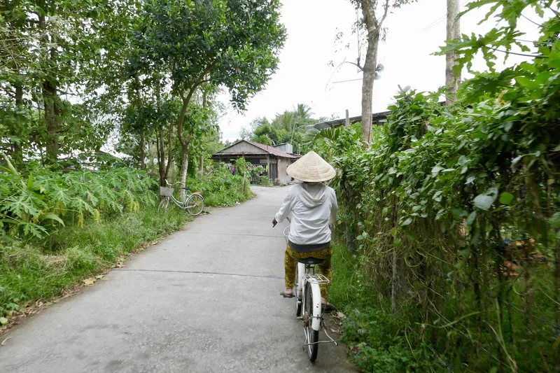 A person in a conical hat cycles along a leafy village path in the Mekong Delta.