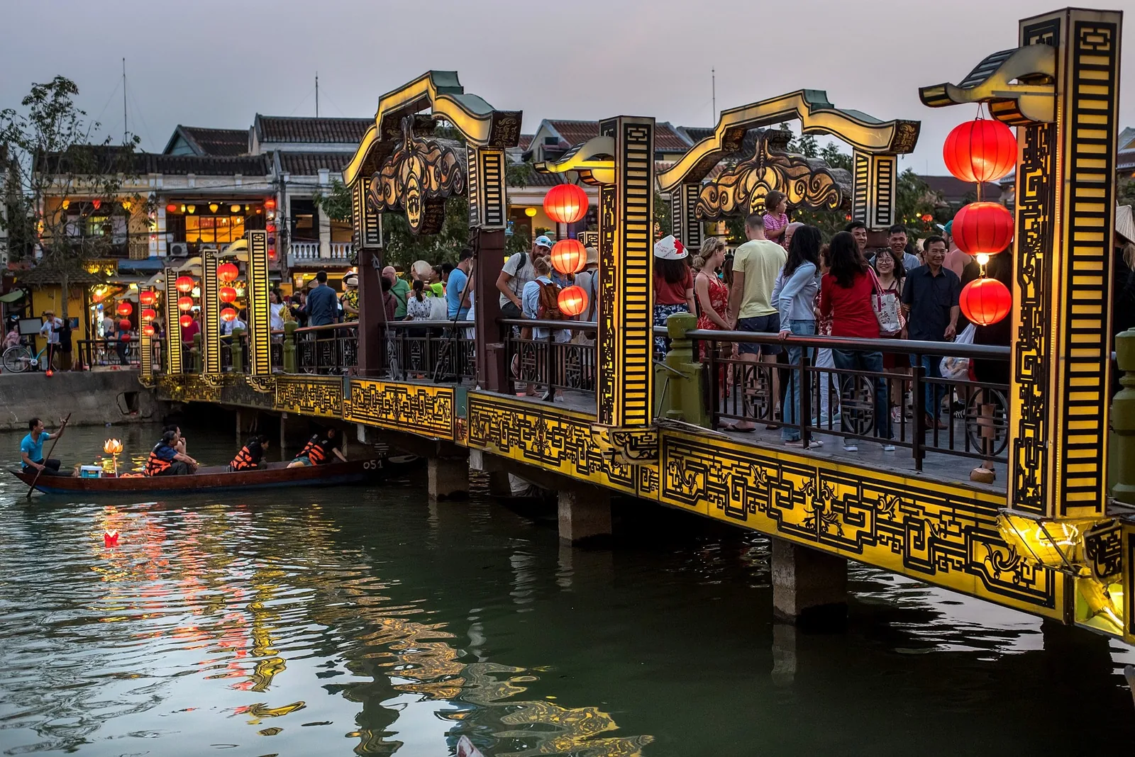 Crowds cross a decorated golden bridge hung with red lanterns above the Hoi An river at dusk.