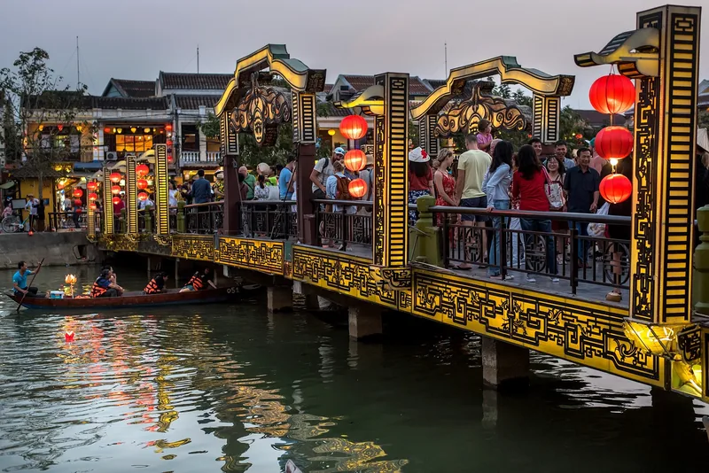 Crowds cross a decorated golden bridge hung with red lanterns above the Hoi An river at dusk.