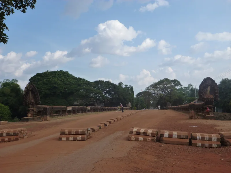 An ancient stone causeway lined with naga balustrades stretches across a dirt road near Siem Reap, Cambodia.