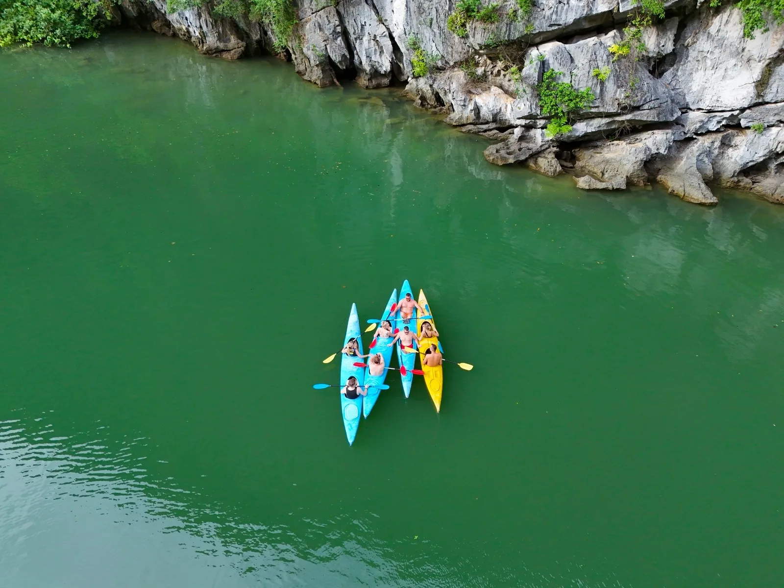 Aerial view of kayakers paddling colorful kayaks on emerald water beside limestone cliffs in Lan Ha Bay.