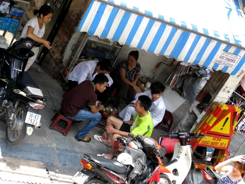 Locals sit on low stools around a small table under a striped awning on a Hanoi sidewalk shop.
