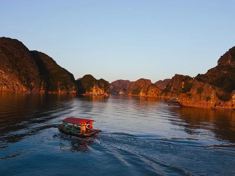 A small red-roofed boat cruises through limestone karsts of Lan Ha Bay at sunset.