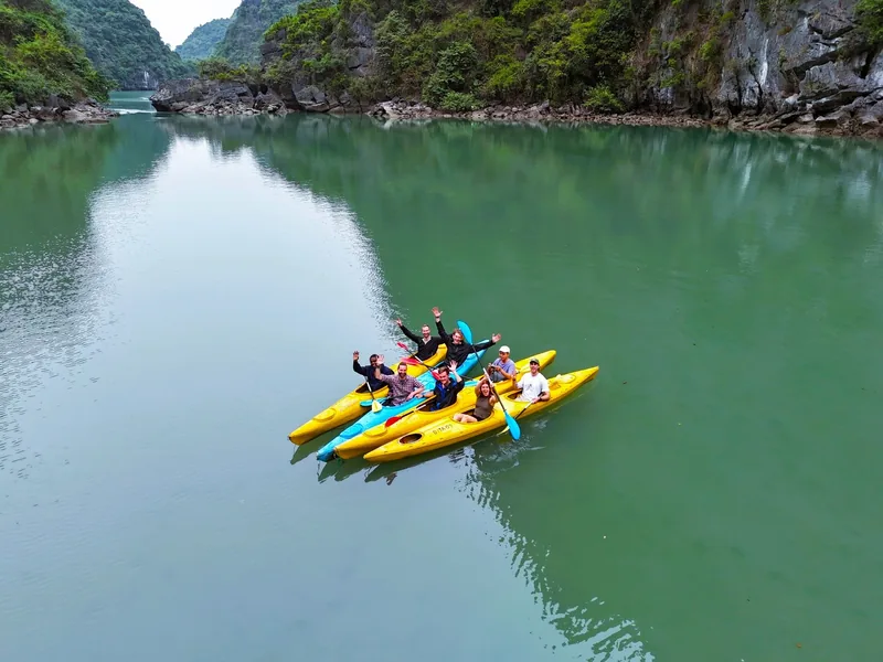 Travelers wave from yellow kayaks on calm emerald water surrounded by limestone karsts in Lan Ha Bay.