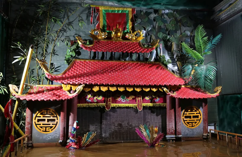 A miniature red-roofed pagoda stage set for a water puppet show in Hanoi, decorated with gold motifs and greenery.
