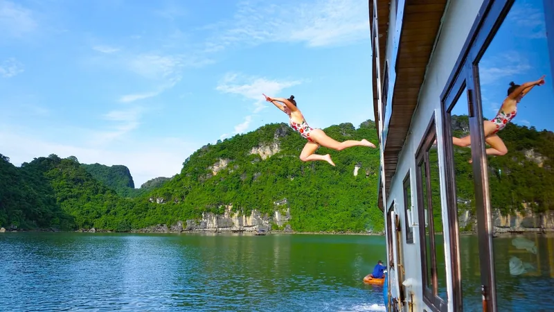 A woman leaps off a cruise boat into the emerald waters of Lan Ha Bay, surrounded by limestone karsts.