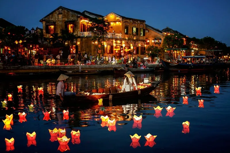 Boatmen in conical hats row past glowing paper lanterns on the river beneath Hoi An's illuminated old town at night.