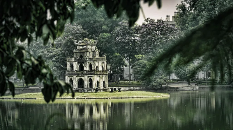 Turtle Tower stands on a small islet in Hoan Kiem Lake, Hanoi, framed by overhanging tree branches.