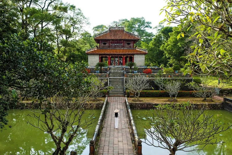 A person in a conical hat walks a brick path over a lotus pond toward the pavilion at Minh Mang Tomb in Hue.