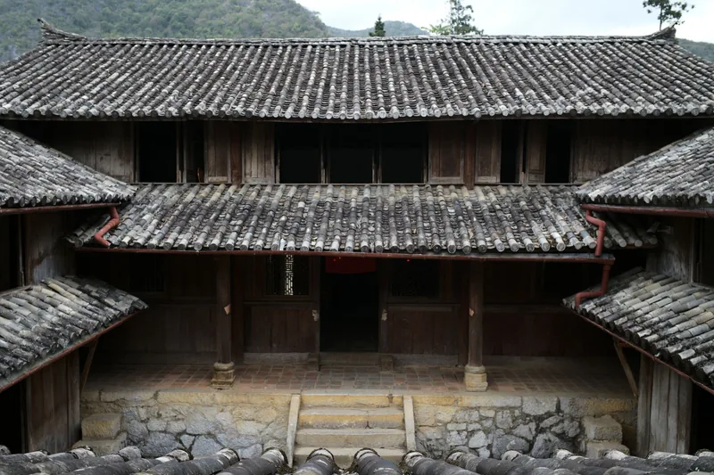 A traditional two-story wooden house with tiled roofs sits below misty mountains in Ha Giang, Vietnam.