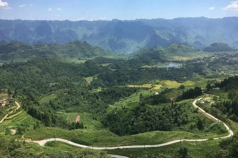 A winding mountain road curves through green valleys and limestone peaks in Ha Giang province.