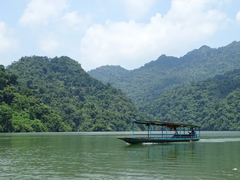 A tourist boat cruises across the calm green waters of Ba Be Lake, surrounded by forested limestone mountains.