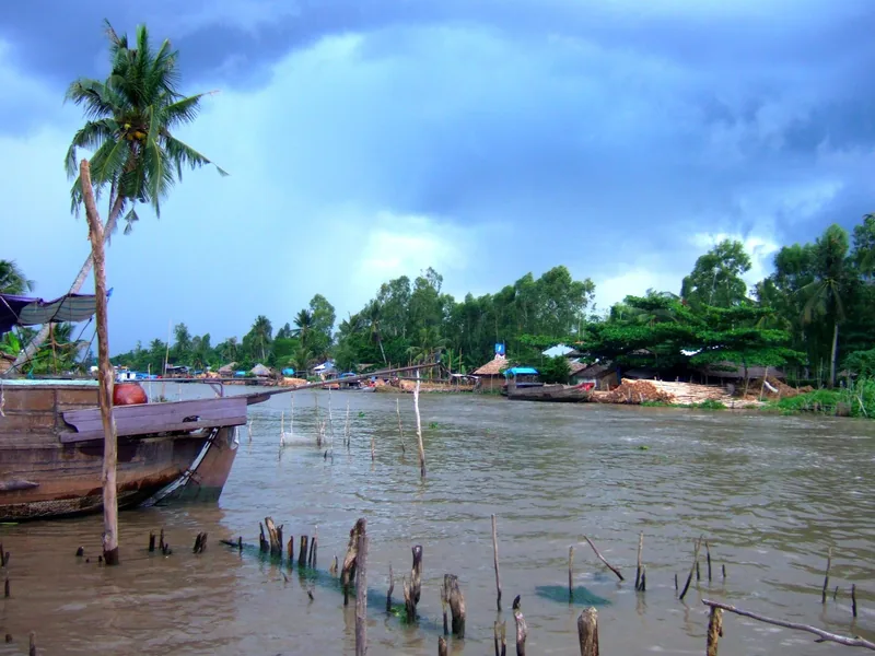 A wooden boat rests on a muddy Mekong Delta river near Soc Trang under a stormy sky.